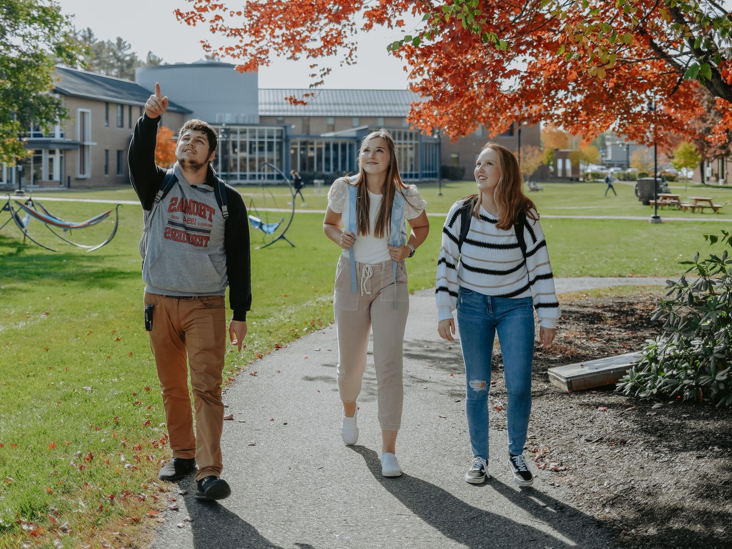 Three students walk on a pathway through campus; one is pointing at something in the distance